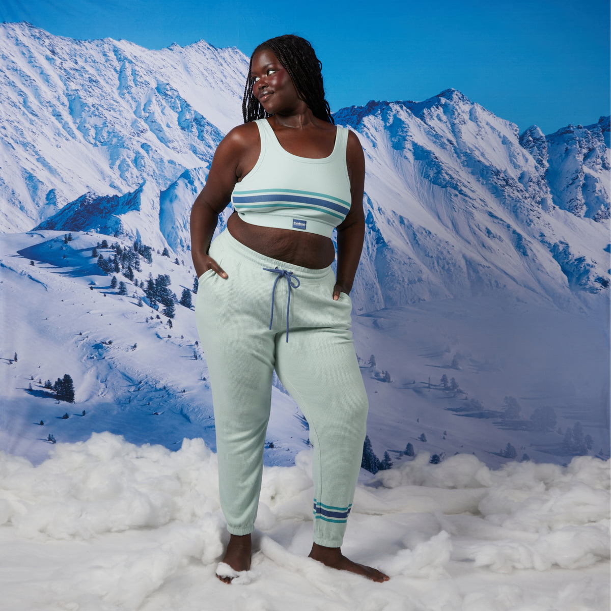 A woman modeling a matching set consisting of an essential soft bra with blue stripes and jogger pants, standing in a snowy mountain landscape backdrop. Model: Rachel is 5'11" and wears a 2X