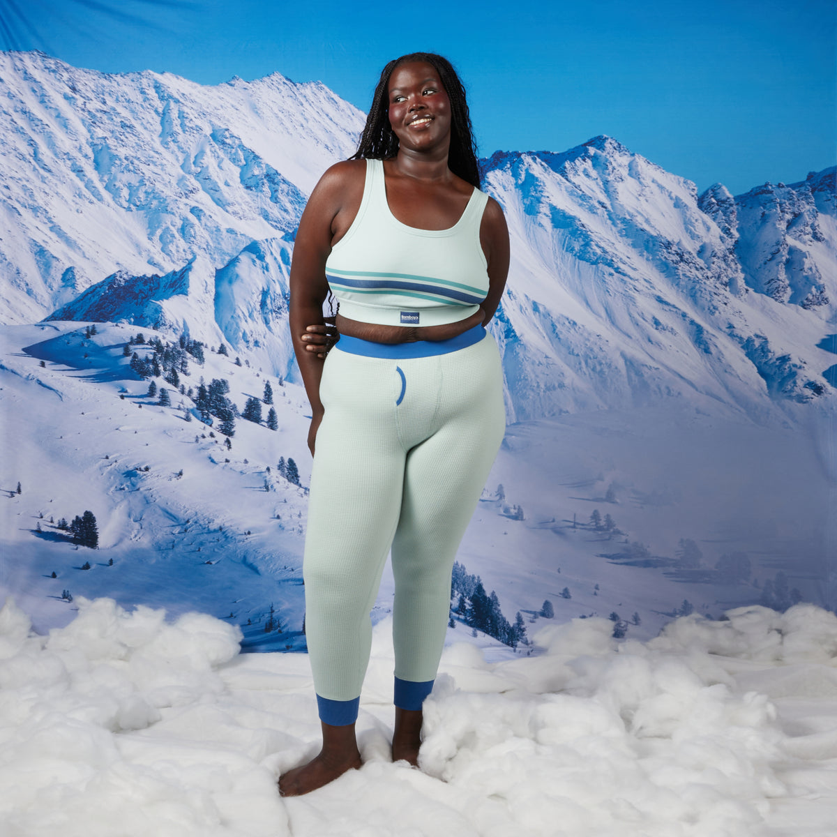 A woman standing in a snowy landscape backdrop wearing a matching light aqua modal cloud waffle set with blue trim, consisting of a essential soft bra and long johns.