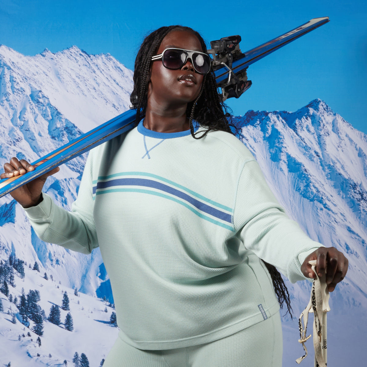 A woman wearing a light aqua and blue striped long-sleeve modal cloud waffle shirt and matching pants, holding skis over her shoulder against a snowy mountain backdrop. Model: Rachel is 5'11" and wears a 2X