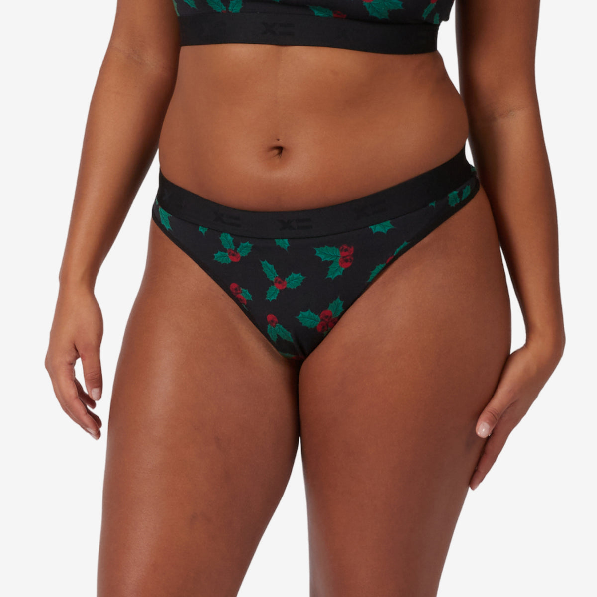 A model wearing black bikini bottoms with a green and red skull holly leaf pattern, shown from the waist to upper thighs against a light gray studio background.