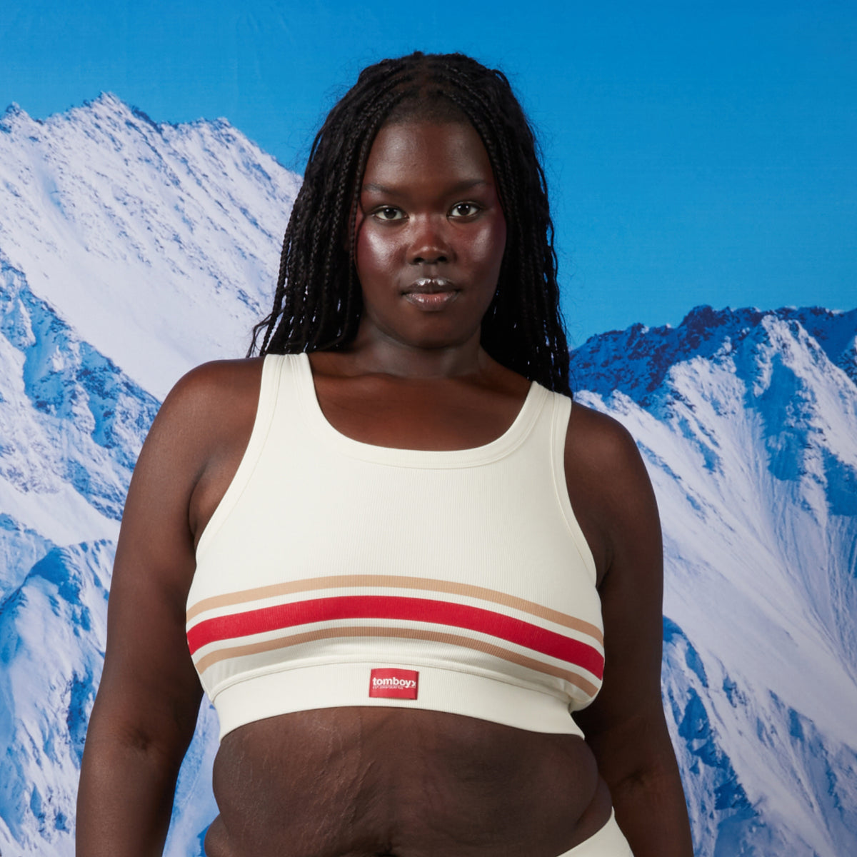 A woman modeling a cream-colored bamboo rib soft bra with red and beige stripes and a small red 'tomboyx' logo. She is facing forward against a backdrop of snowy mountains under a blue sky. Model: Rachel is 5'11" and wears a 2X
