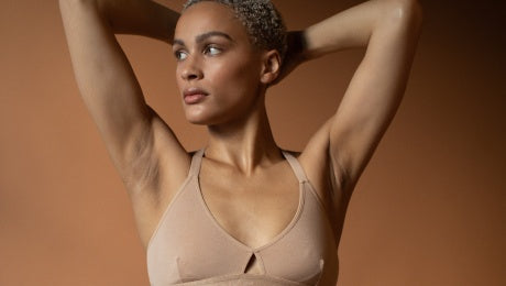 A model looking off to their right, arms above their head, wearing a neutral-toned bralette. 