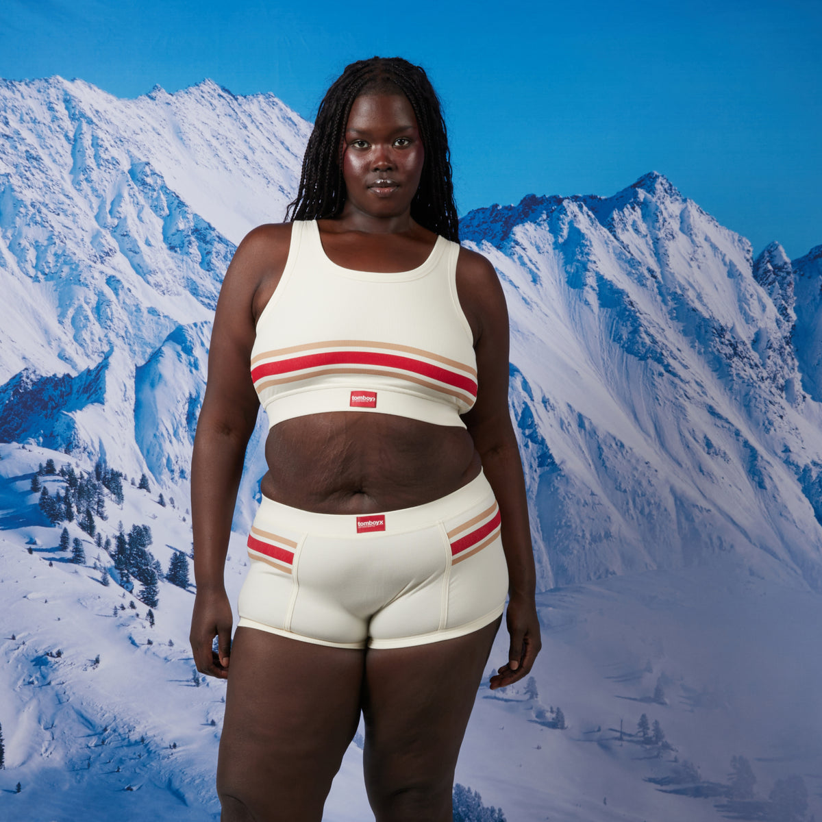 A woman stands facing forward in front of a snowy mountain backdrop wearing a matching cream-colored bamboo rib underwear with red and tan stripes and small red jock tags. The set includes a cropped bra top and boy short style bottoms. Model: Rachel is 5'11" and wears a 2X
